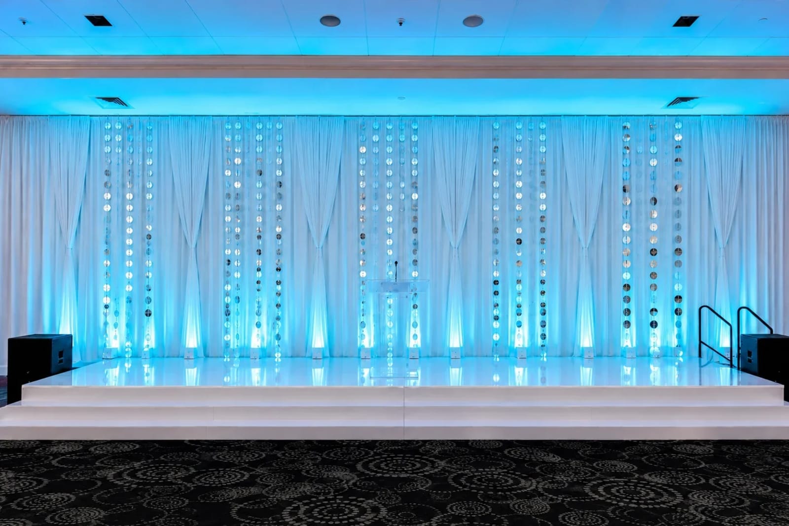 Teal-lit sheer pipe and drape backdrop with a beaded curtain behind a white stage at an Atlanta event.