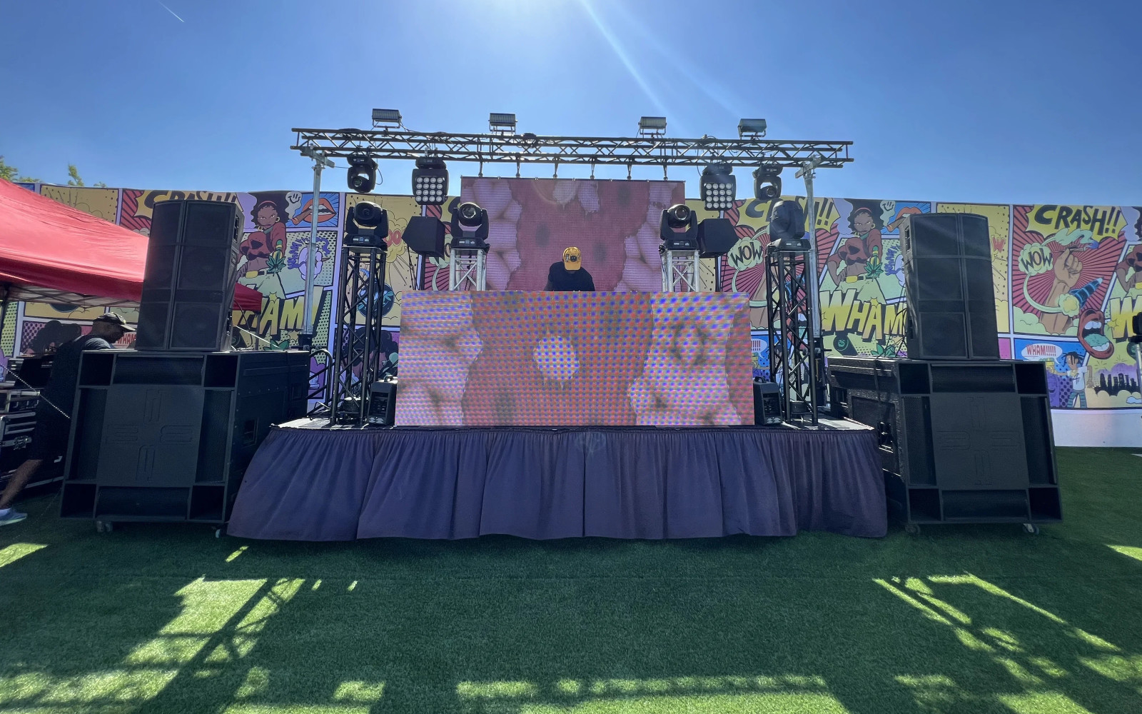 DJ performing on deck stage with LED wall, truss lighting, and subwoofers against pop art backdrop
