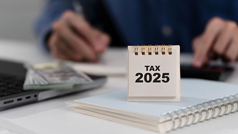 A Tax 2025 calendar on top of a spiral notebook, next to an open laptop with a stack of money sitting on it.