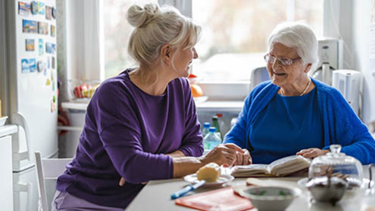 Two older women sitting at a table with a book and food in a kitchen setting.