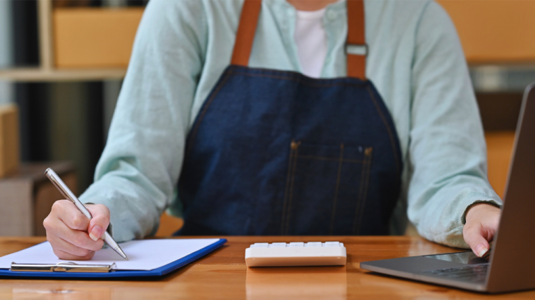 A person in an apron at a desk writing on a clipboard with a calculator in front of them and an open laptop.