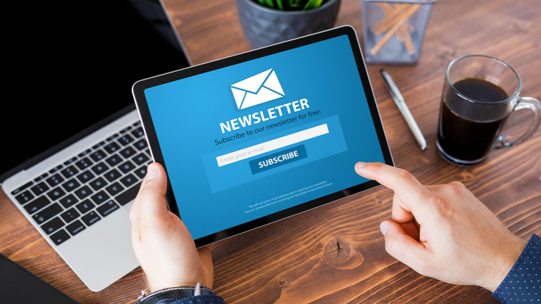 Hands holding a tablet over a desk with a coffee, pen and laptop.