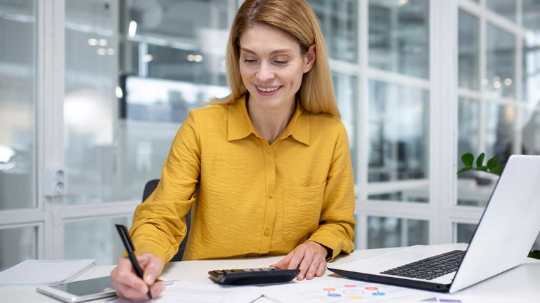 A female business professional sitting at a desk , writing on a clipboard with a laptop, calculator and tablet on her desk.