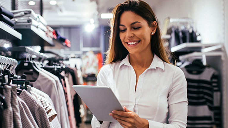 A woman in a clothing store looking at a tablet and smiling.