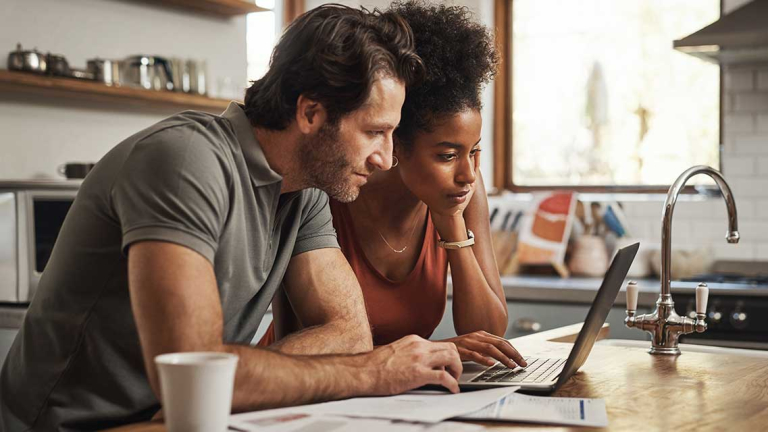 A man and woman leaning on a kitchen counter looking at a laptop with papers scattered around and a coffee cup.