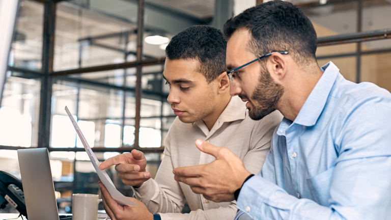 Two men at a desk in an office looking at a sheet of paper with an open laptop, coffee cup and landline phone in front of them.