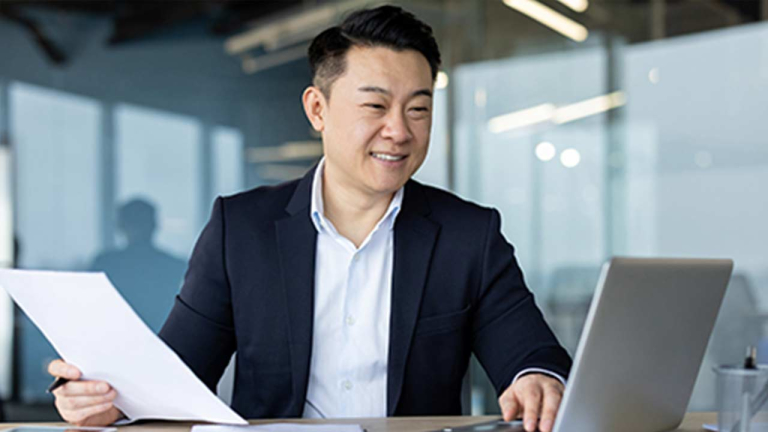 man with paper in hand and typing on laptop
