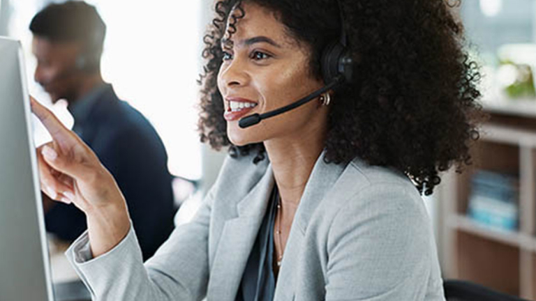 A woman in a headset pointing at a computer monitor.
