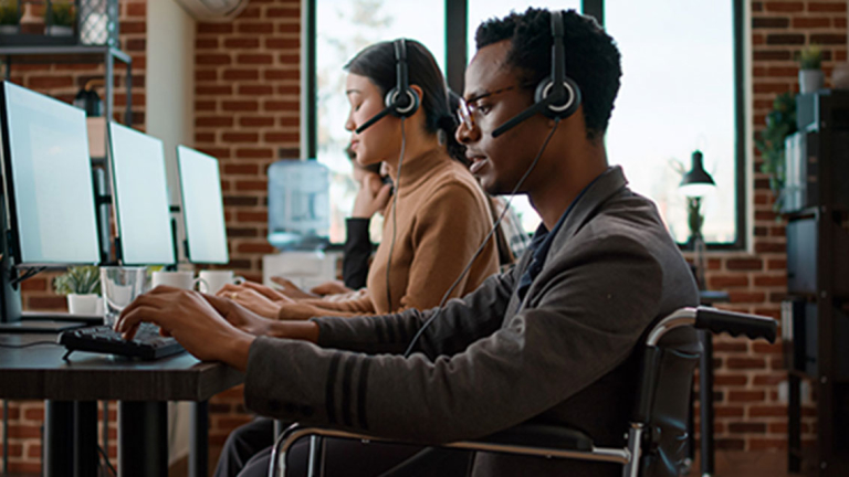 A row of people sitting at a desk, wearing headsets, and using a desktop computer in an office setting.