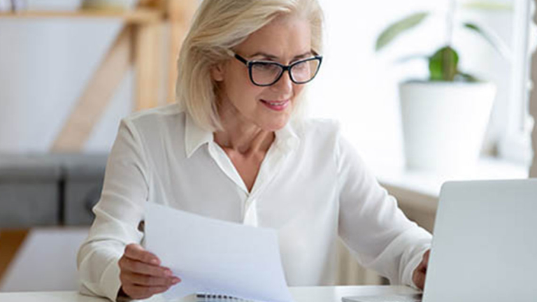 A business woman holding a paper, sitting at a desk using a laptop.