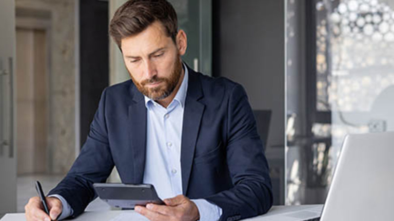 A man in a suit holding a calculator and pen, sitting at an office desk with an open laptop.