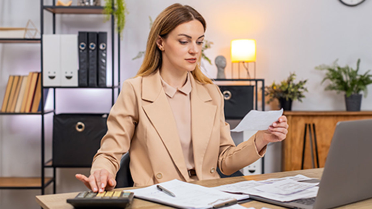A female business professional at a desk, holding a sheet of paper and using a calculator. There are papers, a clipboard, pen, and open laptop on the desk.