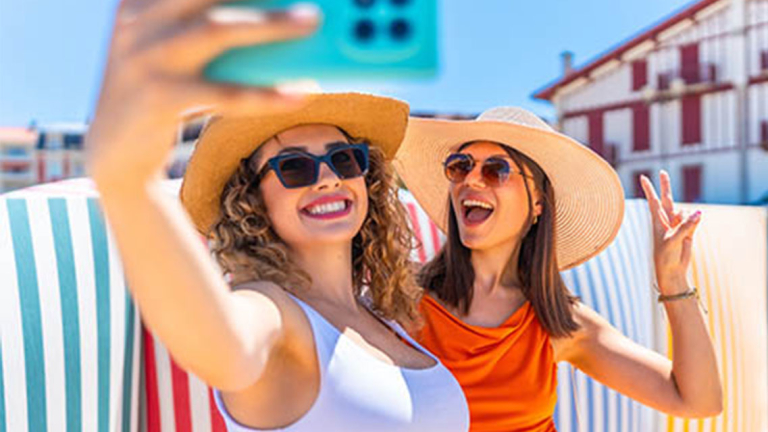 Two women sitting on a beach, posing for a selfie.