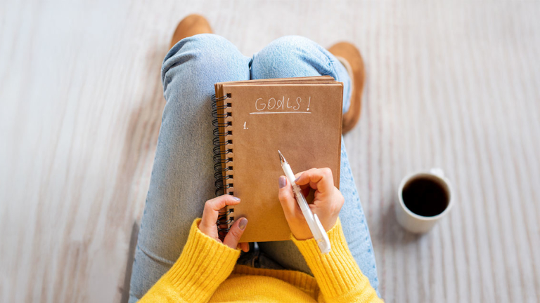 A woman holding a spiral notebook and a pen with the word Goals on it.