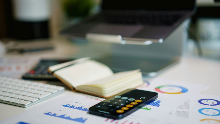 A mobile phone, open notebook, keyboard, calculator and laptop on a desk.