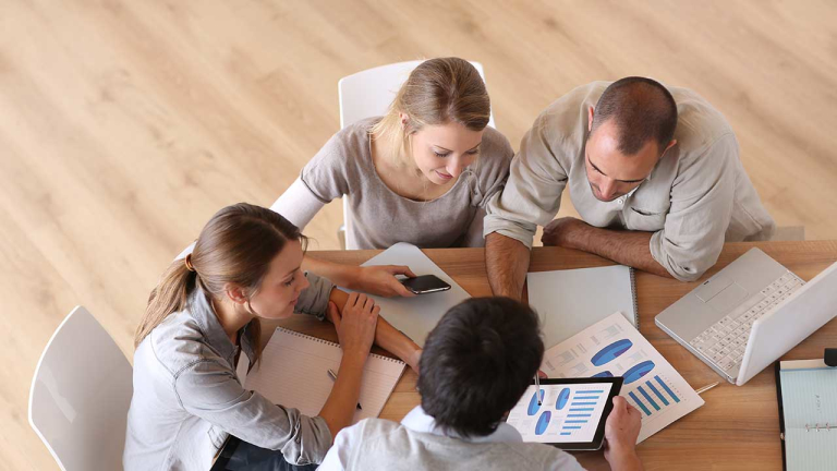 A group of professionals huddled over a desk, looking at graphs on a tablet.