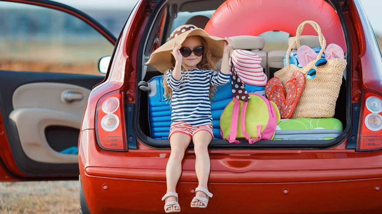 A little girl in a big floppy hat, sun glasses and sandals, sitting in the trunk of a car with beach items.