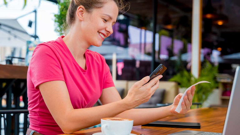 A smiling woman holding a cellphone and receipt while sitting at a table with a coffee and open laptop.
