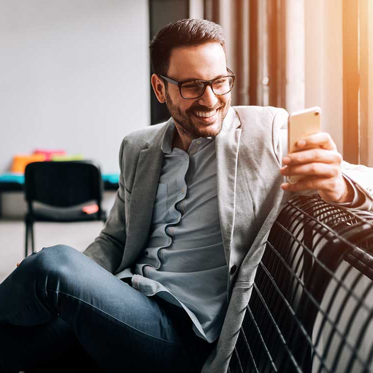 man with glasses wearing sport coat sitting on bench smiling and looking at mobile phone