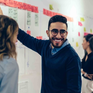 Positive young man laughing while collaborating with colleagues on creating presentation using colorful stickers for productive work in office