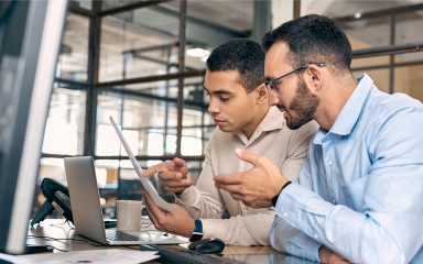 Two men at a desk in an office looking at a sheet of paper with an open laptop, coffee cup and landline phone in front of them.