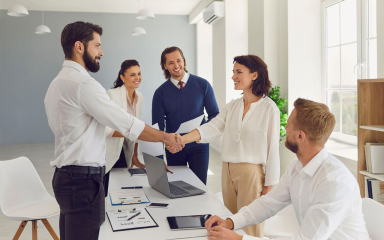 Five business professionals in a meeting room with two of them shaking hands.