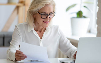 A business woman holding a paper, sitting at a desk using a laptop.