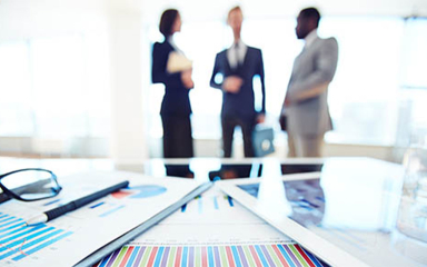 Professionals talking in the background with a tablet, graphs, pen and glasses on a desk.