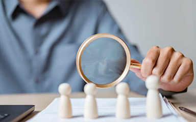 A person holding a magnifying glass looking at wooden figures on a desk with papers and an open laptop.