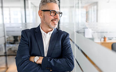 A man in an office building looking through the glass of a meeting area with his arms crossed.