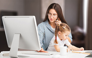 A woman working on a desk top computer, talking on a mobile phone, with a child sitting on her lap, drawing.