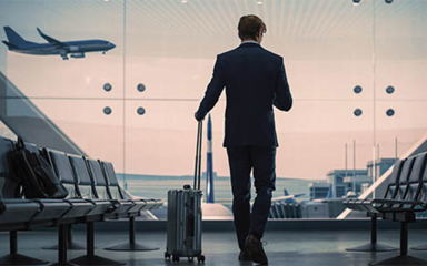 A man in a suit holding rolling luggage and standing in front of a window at the airport with a plane going by.