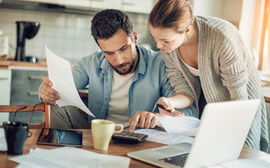A couple at a kitchen table with a calculator, tablet, laptop, papers, and coffee on it.