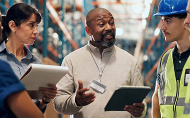 A group of workers, two of which are holding clipboards, talking in a warehouse.