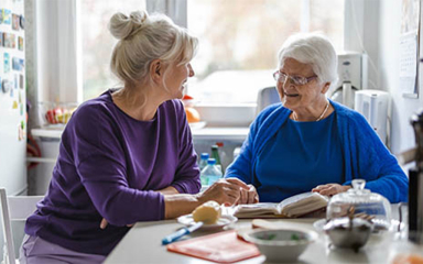 Two older women sitting at a table with a book and food in a kitchen setting.