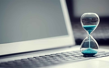Cool toned image of a laptop with a small hourglass on it containing blue sand.