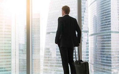 Man with suitcase staring out of a floor to ceiling window in the city.