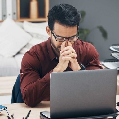 A man sitting at a desk, with an open laptop, and his fingers pushing on the bridge of his nose.