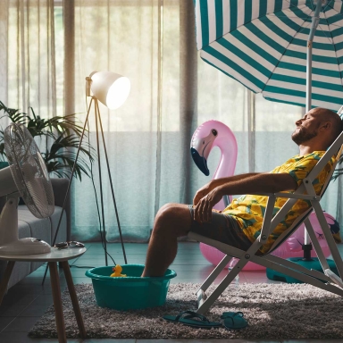 A man sitting in a beach chair with a pink flamingo, umbrella and fan while inside an apartment.