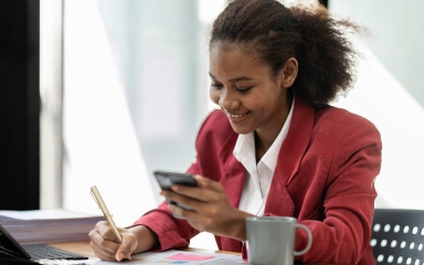 A woman smiling, holding a cellphone and pen, sitting at a desk.