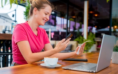 A smiling woman holding a cellphone and receipt while sitting at a table with a coffee and open laptop.
