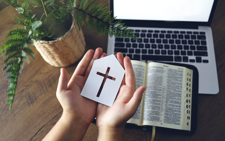 Hands holding a paper cross over a bible and a laptop.