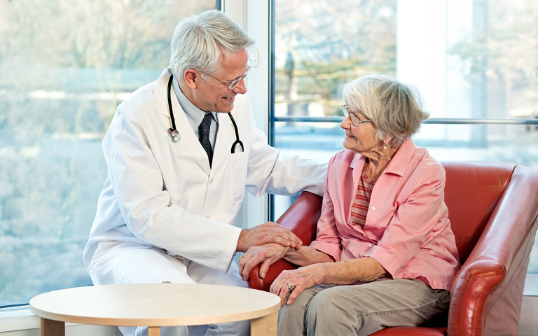 A doctor sitting next to a woman in a chair with one hand on her hand and the other hand on her shoulder.