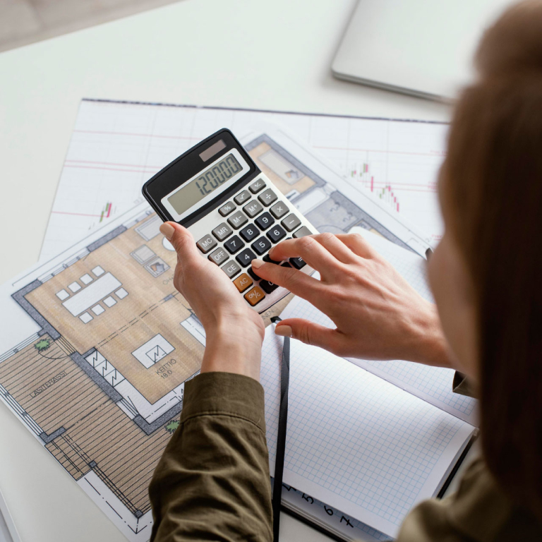 A person using a calculator sitting at a desk with an open notebook and images on papers.