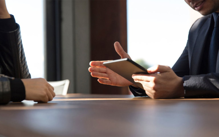 Two people sitting across from one another at a table. One of them is holding a tablet.
