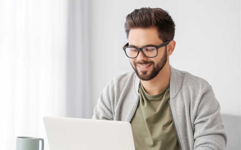 A man in glasses and a hoodie sitting at a desk using a laptop. A cup of coffee in on the desk.