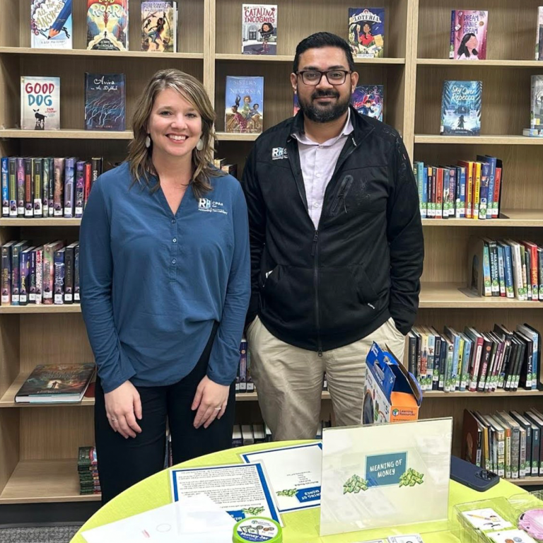 A female and male business professional standing in front of bookshelves with a table in front of them.
