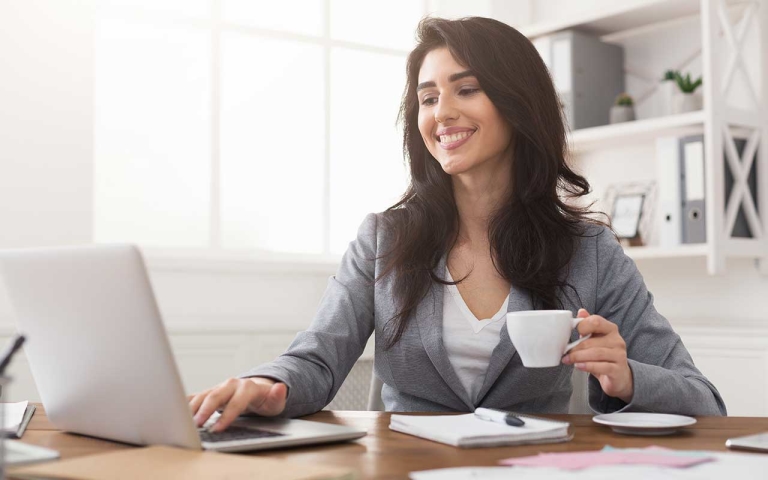 A business woman in an office, using her laptop while holding a cup of coffee and smiling.