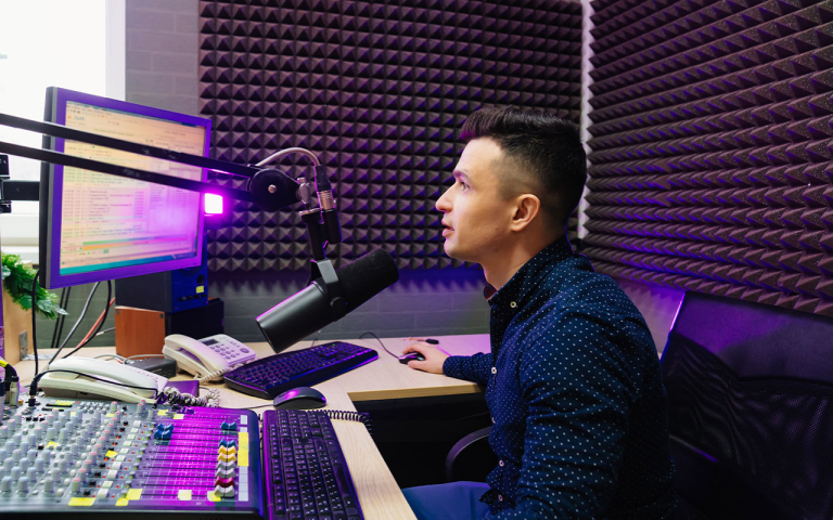 A man looking at a monitor, while talking into a microphone and using a sound board.