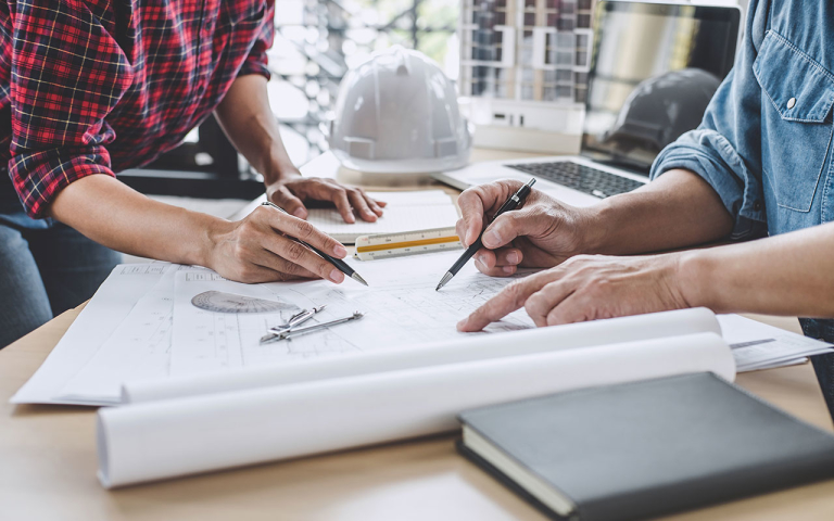 Two people holding pens, drawing on schematics on a table with a laptop, ruler, and hard hat.